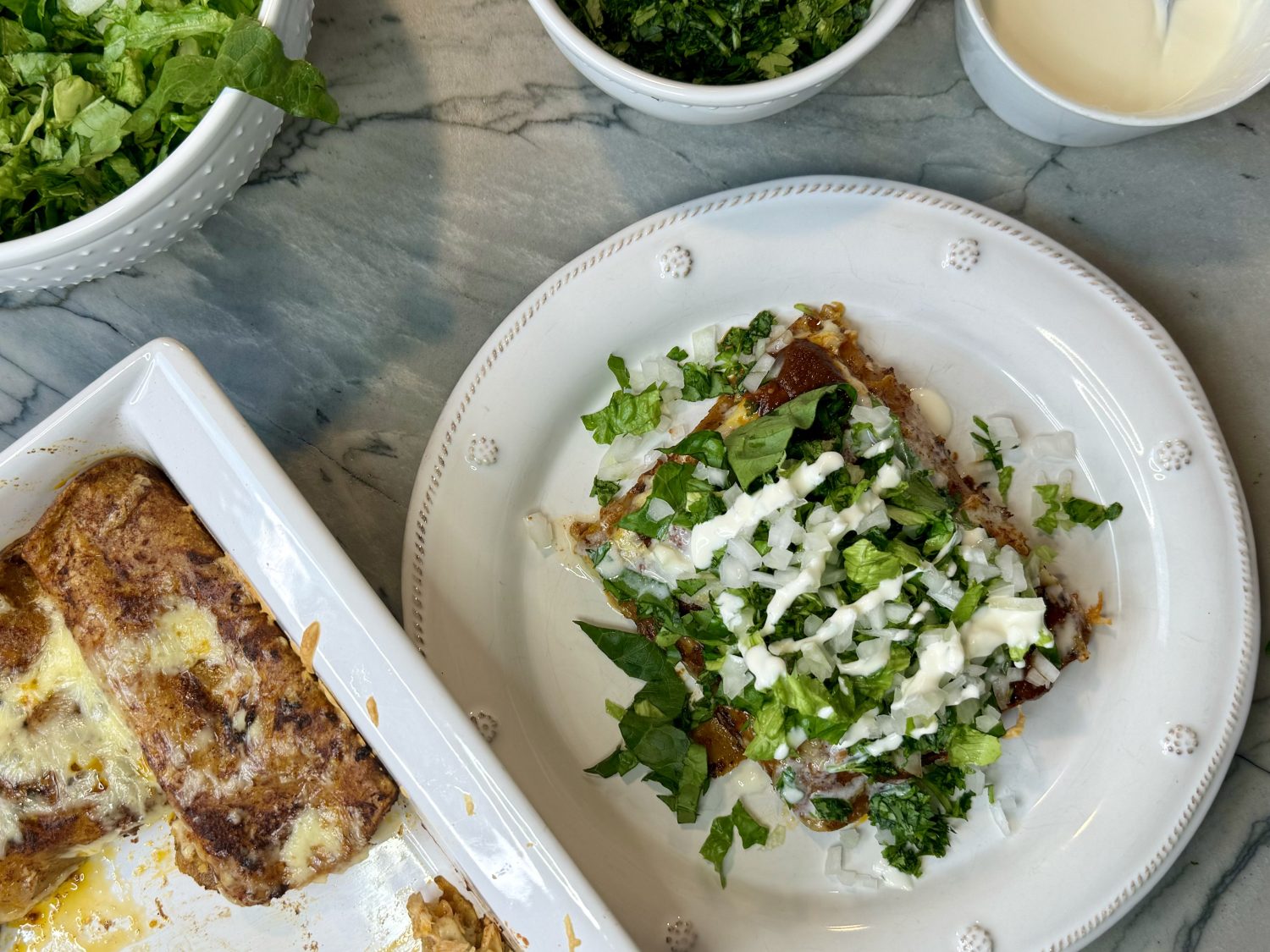 Casserole dish with chicken enchiladas and plate of enchiladas garnished with Chile sauce, lettuce and cream